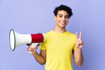 Young Venezuelan man isolated on purple background holding a megaphone and smiling and showing victory sign