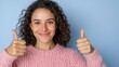 © Iona - A cheerful young woman with curly hair beams with positivity, giving enthusiastic thumbs-ups against a simple blue background.