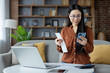 © Liubomir - Asian woman managing personal finances from home with smartphone and laptop. Relaxed atmosphere conveys productivity, focus, and efficiency. Ideal for concepts of remote work and technology.