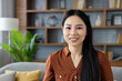 © Liubomir - Asian woman smiling while sitting on sofa in modern home setting. Warm, friendly expression conveys comfort and confidence. Surrounded by wooden bookshelf, plants, creating peaceful ambiance.