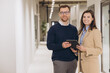 © anatoliycherkas - Smiling business colleagues holding a digital tablet while standing in a modern office hallway, engaging in productive discussion and collaboration