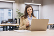 © anatoliycherkas - Smiling businesswoman enjoying a cup of coffee while working on a laptop in a bright, modern office space filled with natural light