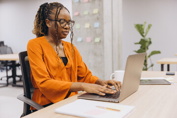 Wall Mural - Focused african american businesswoman typing on laptop, paperwork and greenery nearby, embodying workplace efficiency in contemporary corporate environment