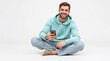 © adynue - smiling happy cheerful young caucasian man sitting cross-legged on the floor wear mint hoody look camera hold in hand use mobile cell phone isolated on plain solid white background studio portrait