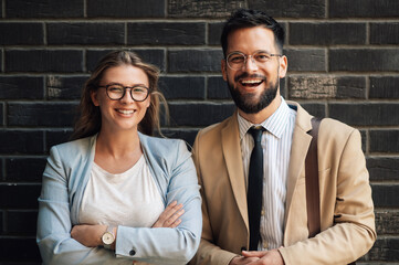 Two business people smiling with arms crossed against brick wall