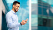 © Prostock-studio - Positive middle eastern guy in formal outfit standing by modern business center, using smartphone and smiling, sending emails while having break, panorama with copy space, side view