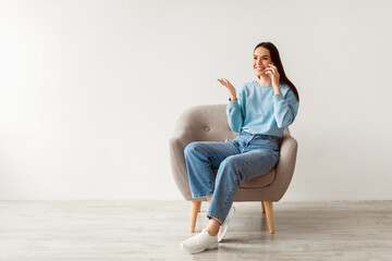 Wall Mural - Mobile call. Cheerful young woman talking on cellphone, sitting in armchair against white wall, empty space for ad. Joyful millennial lady having pleasant phone conversation, enjoying communication