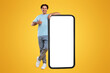 © Prostock-studio - I Recommend. Full body length of cheerful young man leaning on large smartphone with white blank screen, showing thumbs up sign gesture. Gadget with empty free space for mock up, great mobile offer