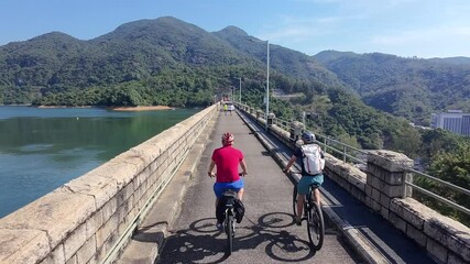 Sticker - cyclists riding on a stone bridge over lake on a sunny day with forested mountain in the background