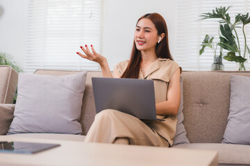 Wall Mural - Woman sitting on sofa with laptop, wearing wireless earbuds, gesturing while smiling in bright living room.