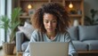 © Janis - Focused young woman with curly hair working at home on laptop, cozy remote work setting