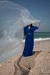 © Milou Dirks - asian young woman in long blue dress on the beach holding fabric in the wind hiding her face