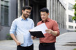 © Liubomir - Two colleagues of office company employees discussing financial reports, contracts and documents, group of business people outside office building with papers in hands discussing.