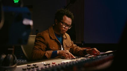 Fototapete - Music producer using audio mixer in professional studio, adjusting volume levels while pressing buttons and sliders on control desk. Sound designer works with digital audio software. Camera A.