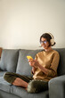© Stock 4 You - Happy smiling young student woman sitting on couch, listening music in headphones on mobile phone application. Relaxing girl at new real estate apartment home using mobile phone online app. Vertical