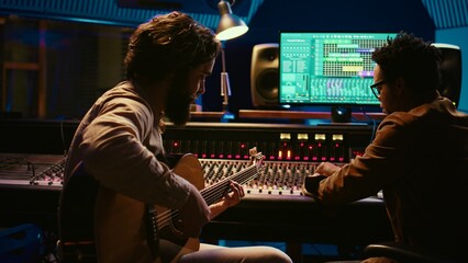 Fototapete - Artist songwriter recording a new track on his electro acoustic guitar in professional studio, using mixing console and gear in control room. Young musician producing new music. Camera B.