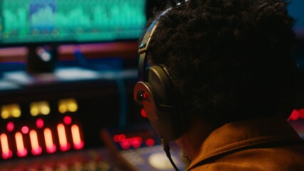 Fototapete - African american tracking engineer processing and mixing sounds in control room, operating audio console with moving faders and colored meters. Technician producer deals with technical gear. Camera A.