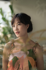 Wall Mural - Asian thoughtful woman in tropical patterned dress gazes into distance, surrounded by soft greenery and natural light