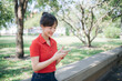 © Jirawatfoto - Asian woman in red shirt smiles while using her smartphone in sunny park, surrounded by greenery and trees