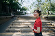 © Jirawatfoto - Asian cheerful woman in red shirt stands outdoors on sunny day, surrounded by trees and steps in park like setting