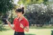 © Jirawatfoto - Asian cheerful woman in red shirt uses her smartphone while standing in sunny park, surrounded by greenery and trees