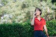 © Jirawatfoto - Asian cheerful woman in red shirt talks on her phone while standing near green hedge in sunny outdoor park