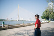 © Jirawatfoto - Asian woman in red shirt smiles while standing near river with bridge in background on sunny day