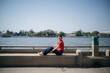 © Jirawatfoto - Asian woman in red shirt and dark pants sits on concrete bench by river, smiling under clear blue sky