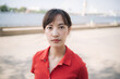© Jirawatfoto - Asian woman red shirt stands outdoors near river, looking directly camera with calm expression. background features blurred view of water and