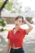 © Jirawatfoto - Asian woman in red cropped shirt and jeans stands outdoors, touching her hair, with thoughtful expression in sunny park setting