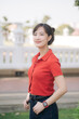 © Jirawatfoto - Asian young woman wearing red shirt and smartwatch stands outdoors, smiling confidently in park setting with blurred background
