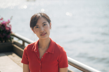 Wall Mural - Asian woman in red shirt stands outdoors near waterfront, with soft sunlight and serene background