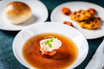 Wall Mural - Soup or consomme, thick meat broth with carrots, chicken dumplings and microgreens, served in a white plate on a dark green background with sesame seed buns, napkin and spoon.