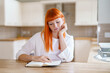 © Iryna - Woman with orange hair writing in a notebook in a bright kitchen during the day