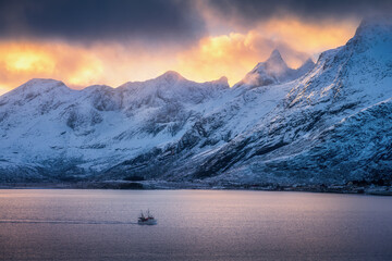 Wall Mural - snowy mountains in clouds, bright sunbeams, boat in fjord at colorful sunset in winter. lofoten isla