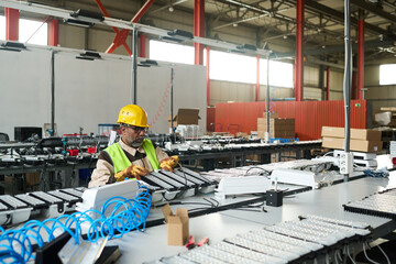 Leinwandbilder - Mature male worker of modern factory producing electric equipment sitting by assembly line and checking new items