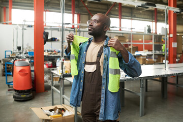 Poster - Young African American worker of warehouse or factory putting on safety vest over denim shirt before working in plant