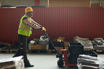 Leinwandbilder - Side view of mature male engineer pushing pallet truck with stack of plastic boxes while moving along aisle in warehouse