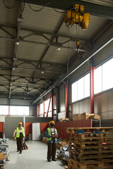 Leinwandbilder - Young engineer in workwear looking upwards and using control pad while his colleague pulling pallet truck in warehouse