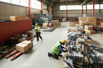 Leinwandbilder - Mature worker pushing pallet truck with packed boxes while passing by young colleague checking packed spare parts