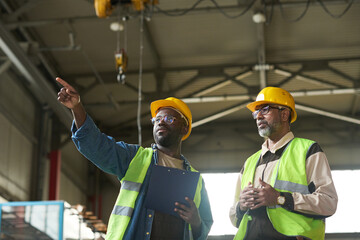 Leinwandbilder - Mature foreman standing next to young male subordinate pointing at new industrial equipment during discussion of its quality