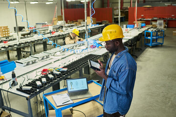 Poster - Serious young African American engineer in safety helmet looking at lamp produced by their factory while standing in workshop