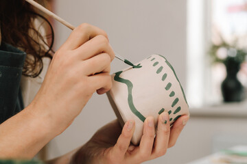Poster - Female artist is painting earthenware with paintbrush and water colors in the workshop. Painter is drawing and making a colorful decoration of a clay ware.