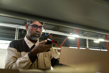 Leinwandbilder - Mature multiethnic factory worker scanning barcodes on packed cardboard boxes while making revision in warehouse