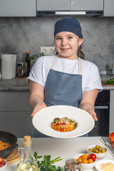 Wall Mural - Portrait of a beautiful little girl in a gray bandana and apron holding a plate of pasta in her hands. The girl shows the spaghetti she has prepared. Close-up.