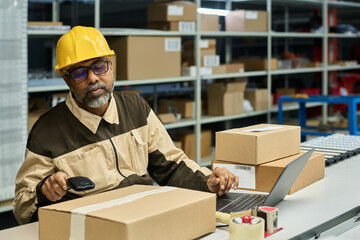 Leinwandbilder - Bearded mature male engineer in safety helmet and workwear sitting by workplace and scanning barcode on sticker on box