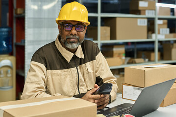 Leinwandbilder - Happy multiethnic warehouse worker in hardhat and workwear looking at camera while scanning barcodes by workplace
