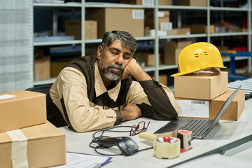 Leinwandbilder - Tired warehouse worker napping in front of laptop screen by workplace among packed cardboard boxes after revision
