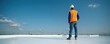 © miss irine - Roofer in orange vest, protective hard hat stands on roof coated with reflective paint. Man inspects flat commercial roof for energy efficiency, renovation. Contractor looks away at clear blue sky.