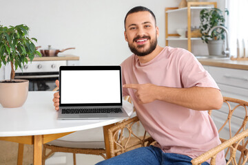 Young man pointing at blank laptop on table in kitchen